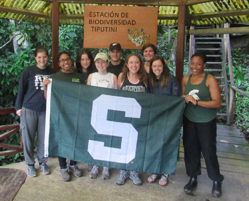 a diverse group of education abroad students in Ecuador, holding a green Spartan S flag, smiling at the camera. They are on a wooden platform with thick foliage behind them, and a sign that reads, in Spanish, Estación de Biodiversidad Tiputini